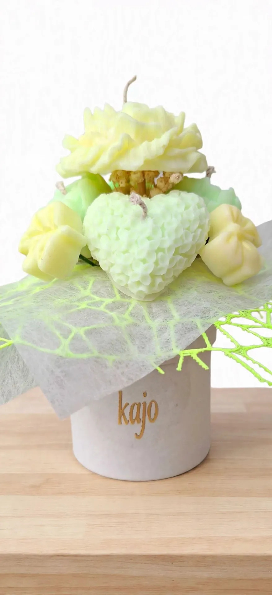 Vaaleankeltainen kukka-asetelma valkoisessa ruukussa, jossa teksti “kajo”, taustalla vihreä koristeverkko.
